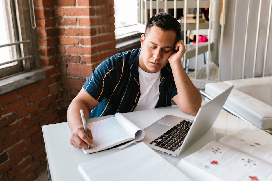 A young man falls asleep while studying at a desk with a laptop.