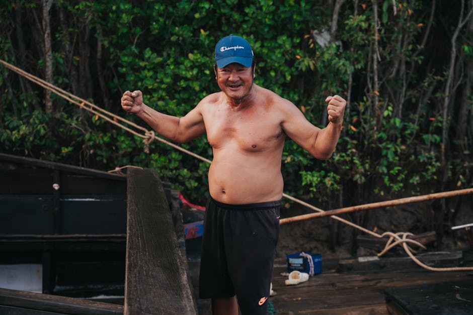 A cheerful shirtless man flexes his muscles outdoors, wearing a blue cap.