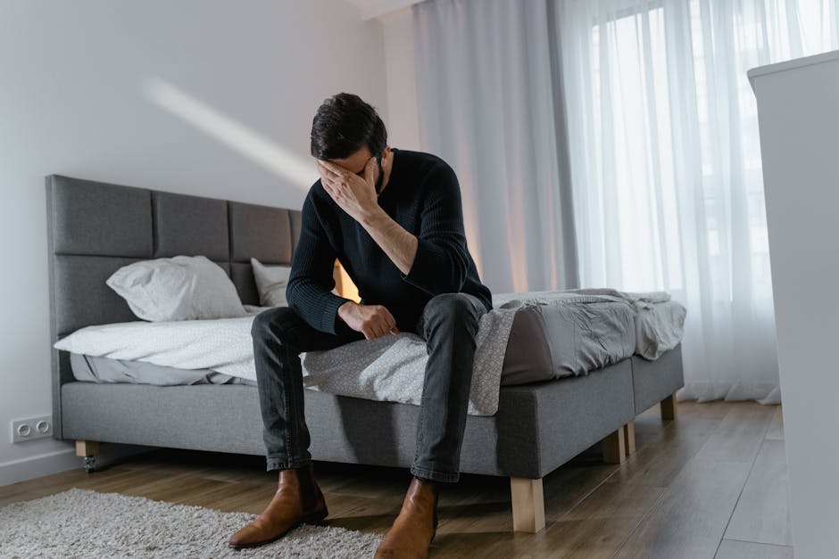 A thoughtful man in a black sweater sits on a bed in a modern bedroom.