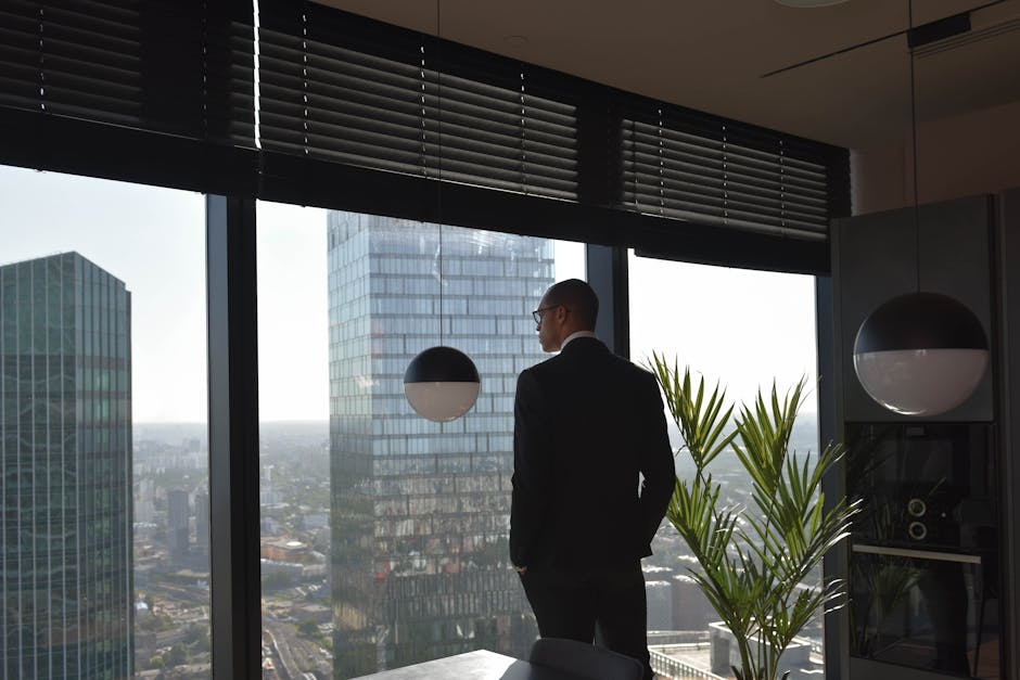 A businessman in a black suit looks out at skyscrapers from an office window, embracing an urban view.