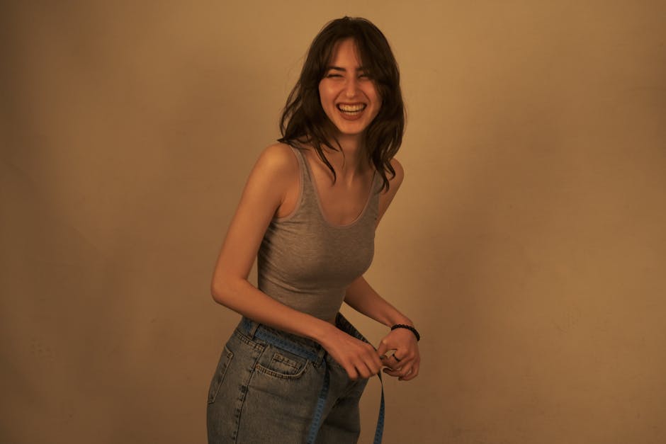 Cheerful woman in casual attire measuring waist with a tape measure indoors.