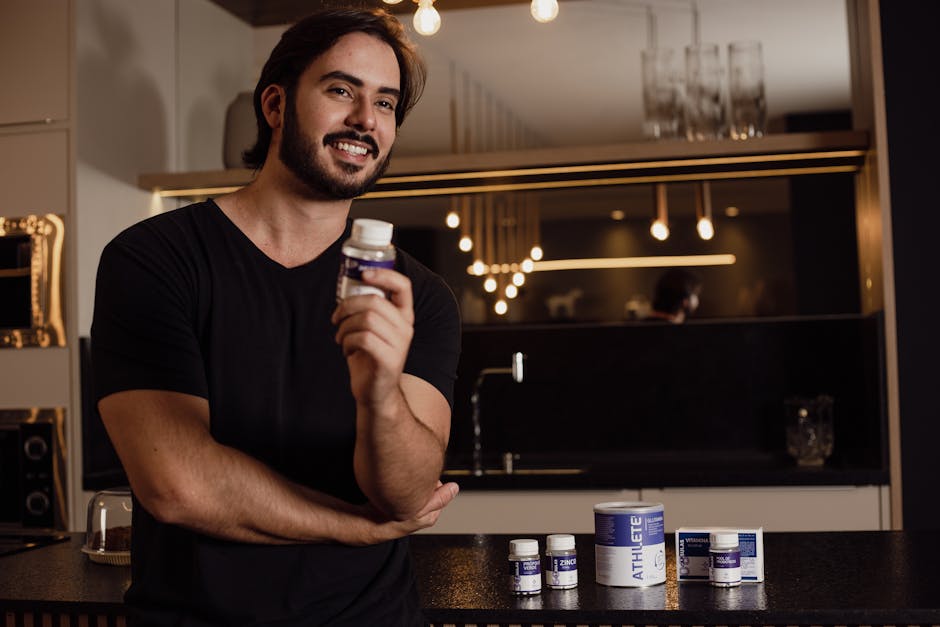 Smiling man holding a supplement bottle in an indoor kitchen setting with warm lighting.