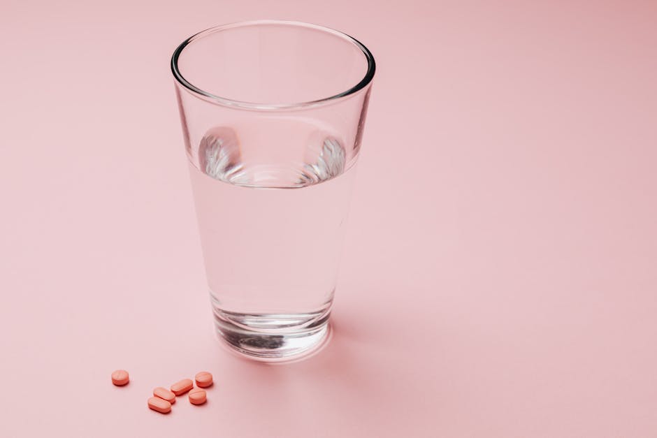 Glass of water with scattered pink pills on a pastel pink background, symbolizing medication or daily supplements.
