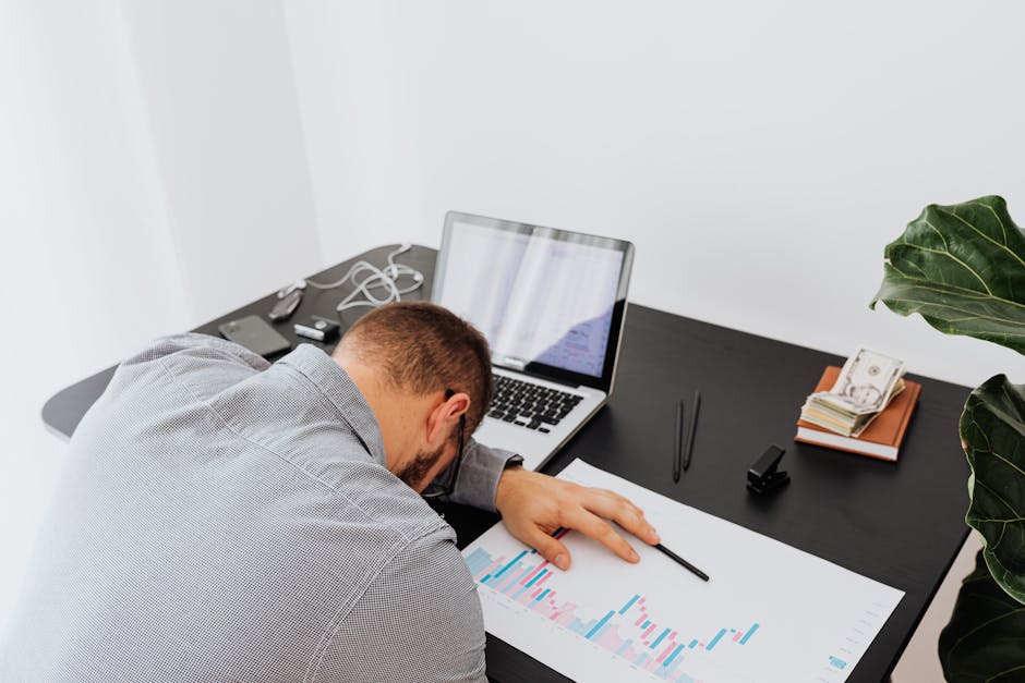 Overworked businessman falling asleep on office desk with laptop and charts, depicting stress and exhaustion.