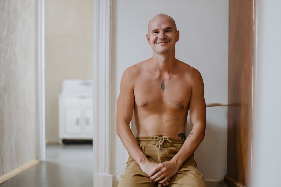 Bald tattooed man sitting shirtless indoors, smiling warmly.
