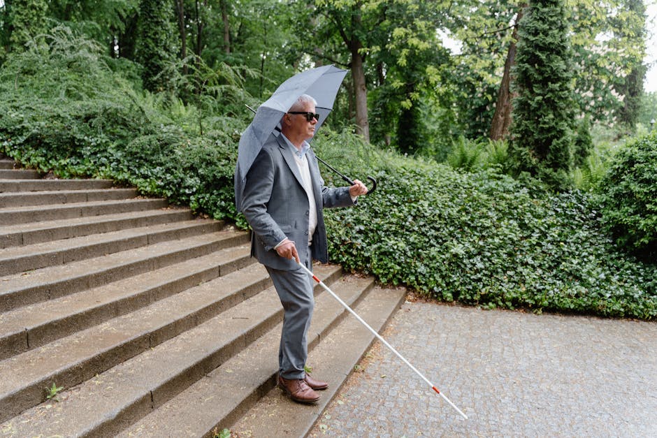 Senior man with visual impairment walks with white cane and umbrella in a green park.