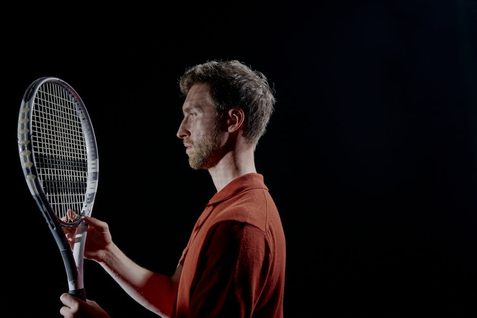 Profile view of a focused male tennis player holding a racket in a dimly lit setting.