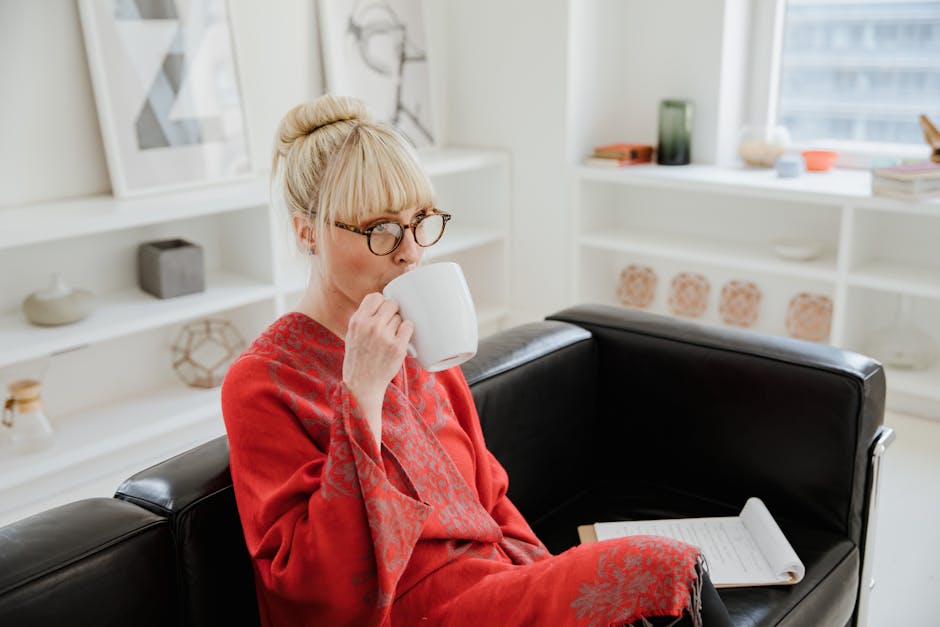 A woman in a red bathrobe drinks coffee while sitting on a sofa indoors.