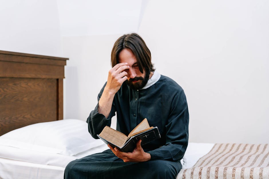 A man in a soutane sits on a bed, deeply engaged in prayer while reading the Bible.