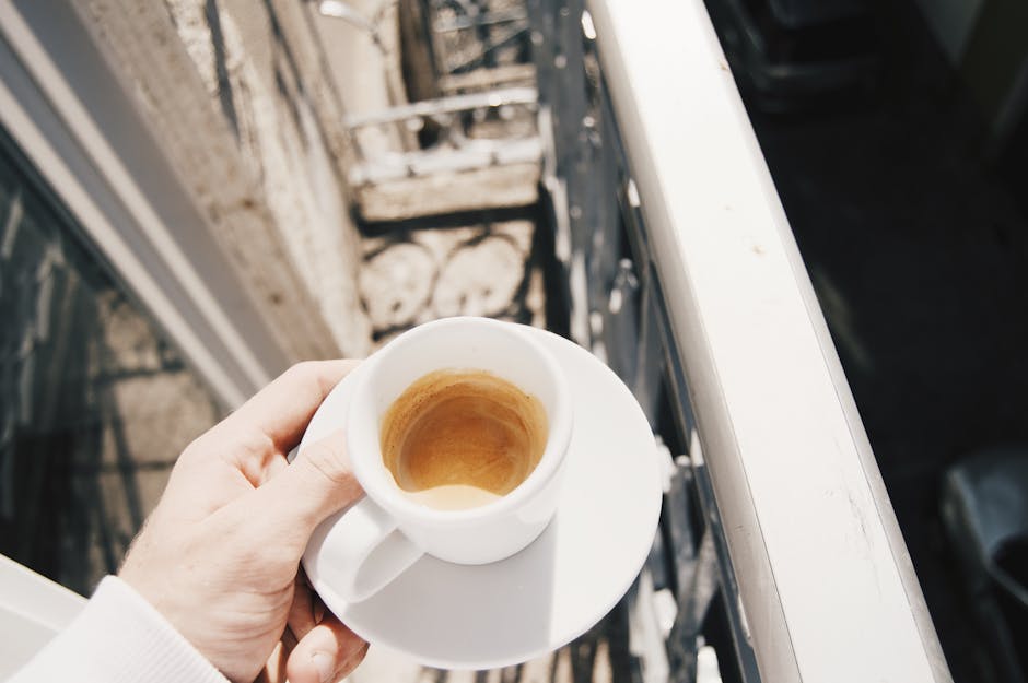 From above of anonymous man holding saucer with cup of leftover cappuccino coffee on small balcony in city on sunny day