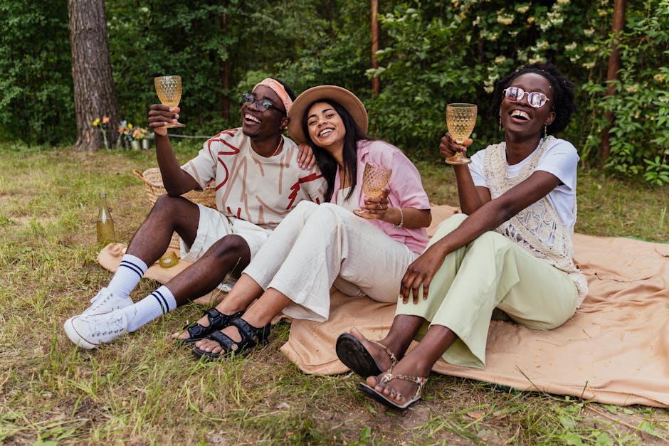 Three friends laughing and enjoying a picnic outdoors with drinks.