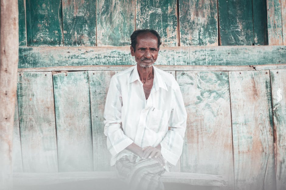 Elderly man sitting against a rustic wooden backdrop in Mandya, India.
