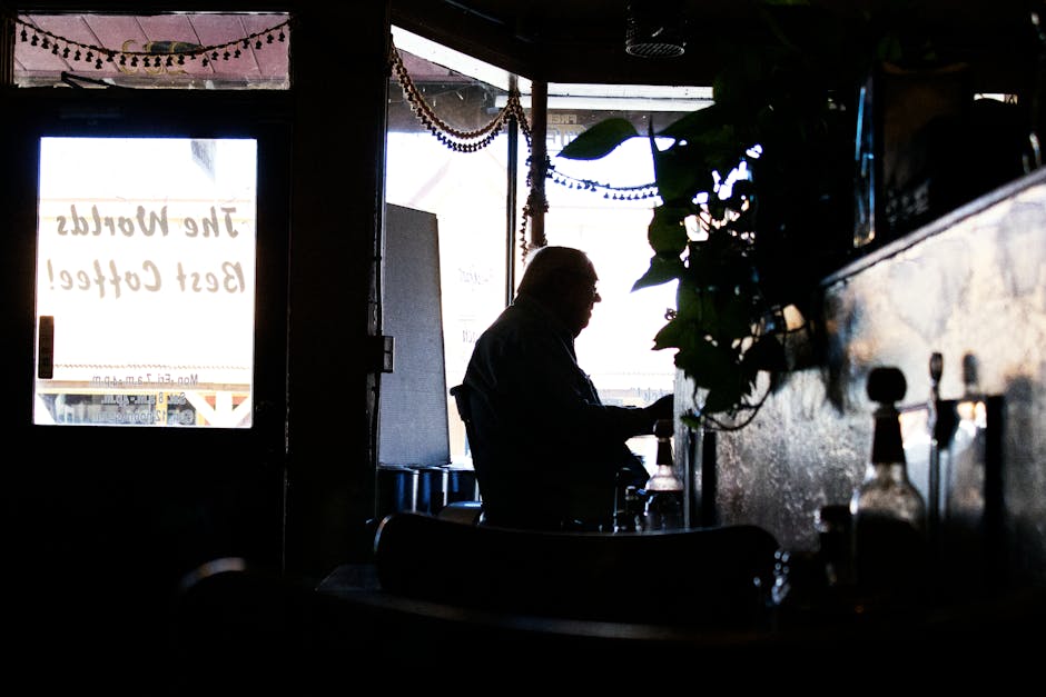 A silhouette of an elderly man sitting in a dimly lit coffee shop, surrounded by plants.
